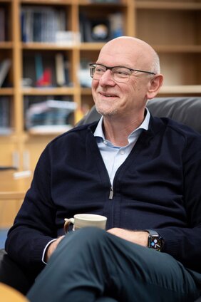 Professor Dr. Martin Winter sitzt in blauer Strickjacke in einem Sessel und hält eine Tasse in den Händen.