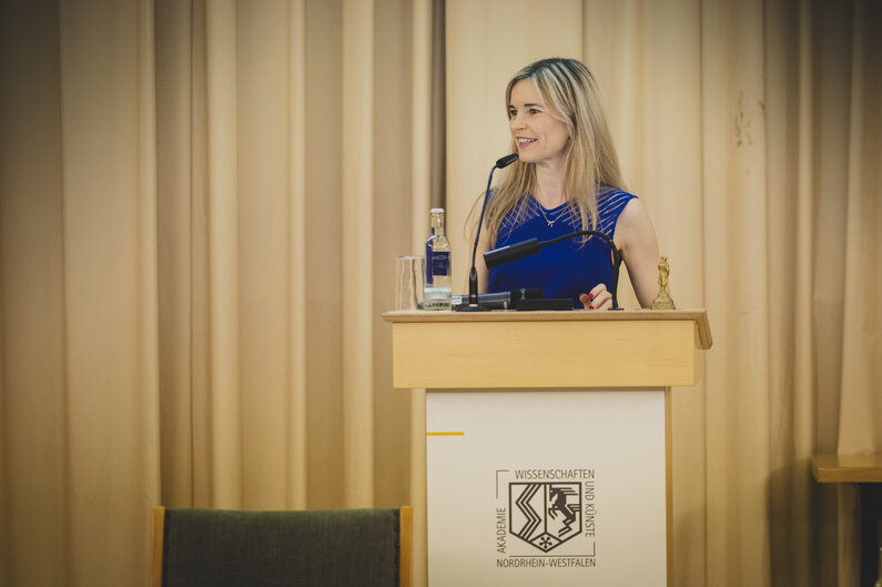 Eine Frau mit blonden langen Haaren und einem blauen Kleid steht an einem Rednerpult.