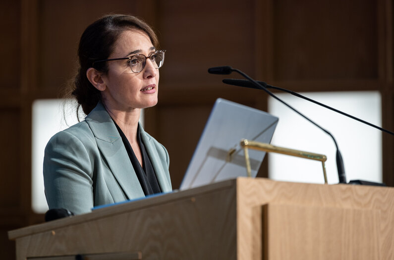 Eine Frau steht an einem Rednerpult. Sie spricht in ein Mikrofon.