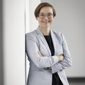 Eine Frau lehnt an einer weißen Wand. Sie lächelt in die Kamera.