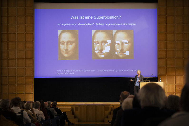 Eine Frau steht neben einem Rednerpult. Auf dem Bildschirm hinter ihr sind drei Fotos der Mona Lisa zu sehen. 