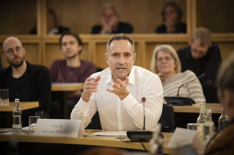 Ein Mann sitzt an einem Tisch. Er gestikuliert mit den Händen. Auf einem Schild steht sein Name "Prof. Dr. Andreas Nitschke".