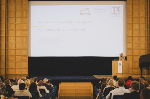 Eine Frau steht auf einer Bühne. Im Hintergrund ist der Titel der Veranstaltung eingeblendet "Pierre Boulez in Duisburg-Marxloh.