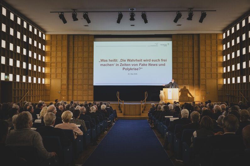 Ein Saal voller Menschen. Auf der Bühne steht ein Mann hinter einem Rednerpult.