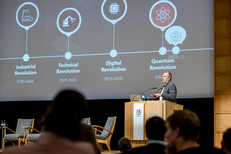 Ein Mann steht hinter einem Rednerpult. Im Hintergrund sind verschiedene technische Epochen eingeblendet. 