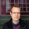 Ein Mann mit Brille steht lächelnd vor einem Fenster.