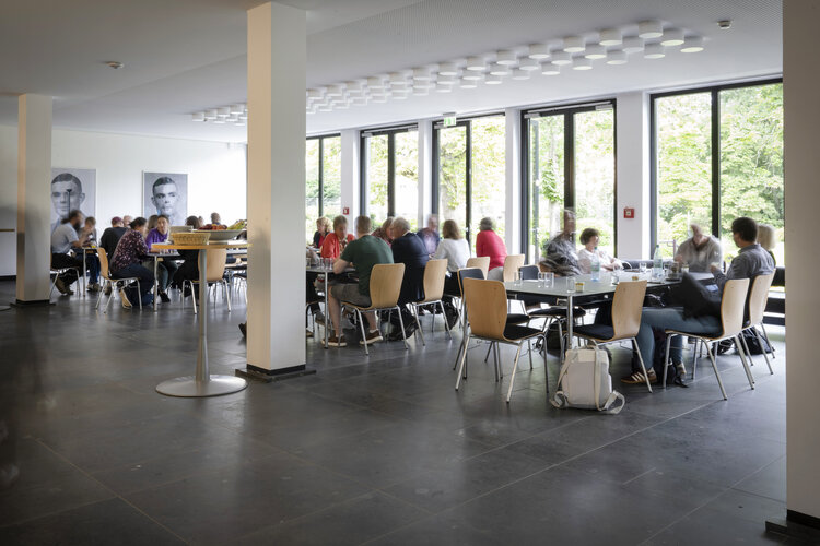 Männer und Frauen sitzen an großen Tischen im Foyer der Akademie. 