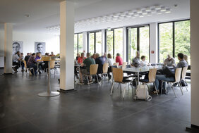 Männer und Frauen sitzen an großen Tischen im Foyer der Akademie. 