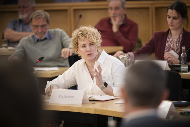 Eine Frau sitzt an einem Tisch. Auf einem Schild steht ihr Name "Kristin Küter".