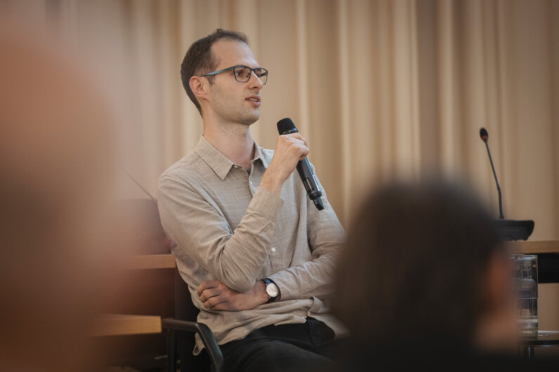 Ein Mann sitzt in einem Vortragsraum. In der Hand hält er ein Mikrofon.