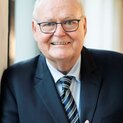 Ein Mann mit Brille lehnt an einer Wand. Er lächelt in die Kamera und hält seine rechte Hand auf Brusthöhe.
