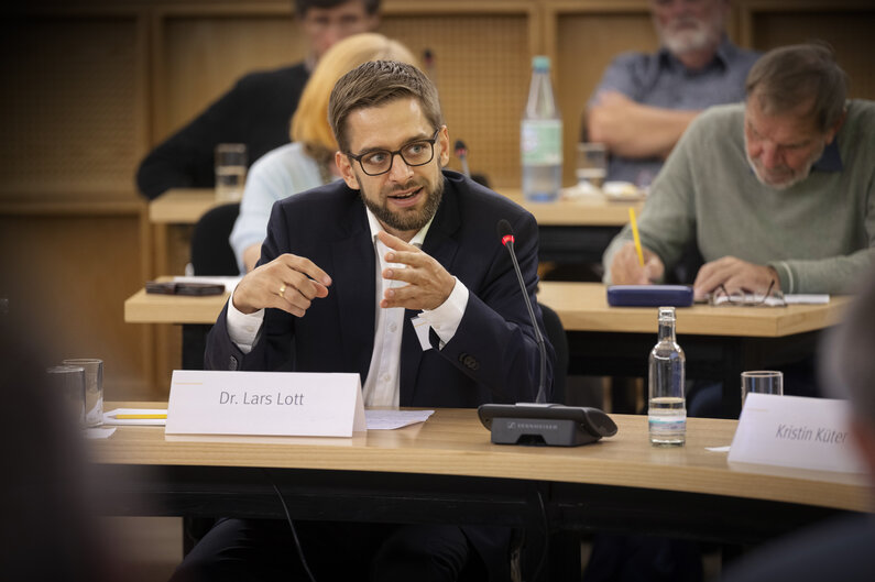 Ein Mann sitzt an einem Tisch. Auf einem Schild steht sein Name "Dr. Lars Lott".