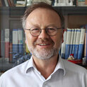 Ein Mann mit Brille steht vor einem Bücherregal und lächelt.