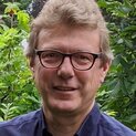 Ein Mann mit Brille steht vor einem Strauch und lächelt in die Kamera.