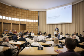 Menschen sitzen in einem Vortragsraum an Tischen. Ein Mann steht hinter einem Rednerpult.
