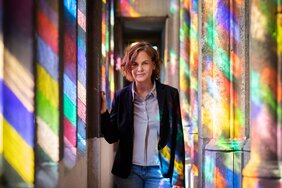 Das Foto zeigt ein Porträt der Dokumentarfilmerin Corinna Belz. Sie steht im Triforium des Kölner Doms vor dem Südquerhausfenster. An den Wänden spiegelt sich die farbige Verglasung des Fensters.