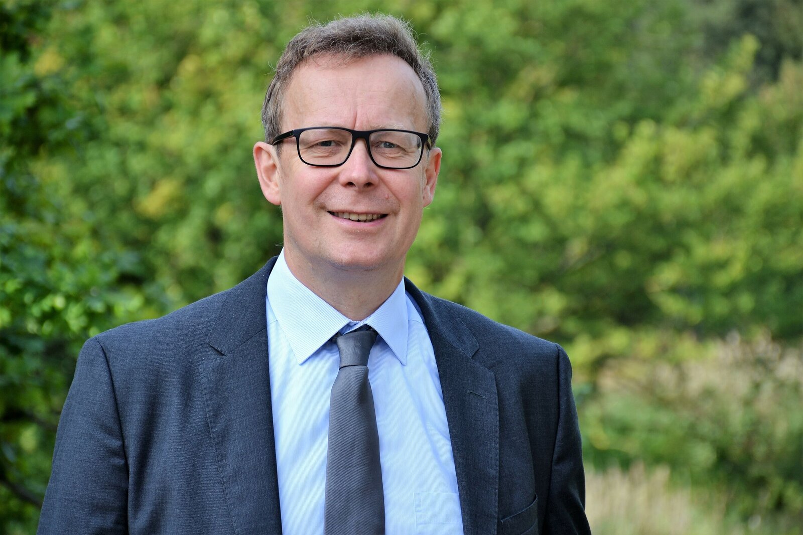 Ein Mann mit Brille steht lächelnd vor Sträuchern. Er trägt eine Anzugjacke und Krawatte.