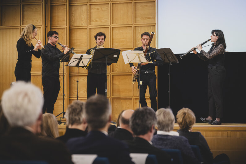 Zwei Musikerinnen und drei Musiker stehen auf einer Bühne. Sie spielen verschiedene Instrumente.