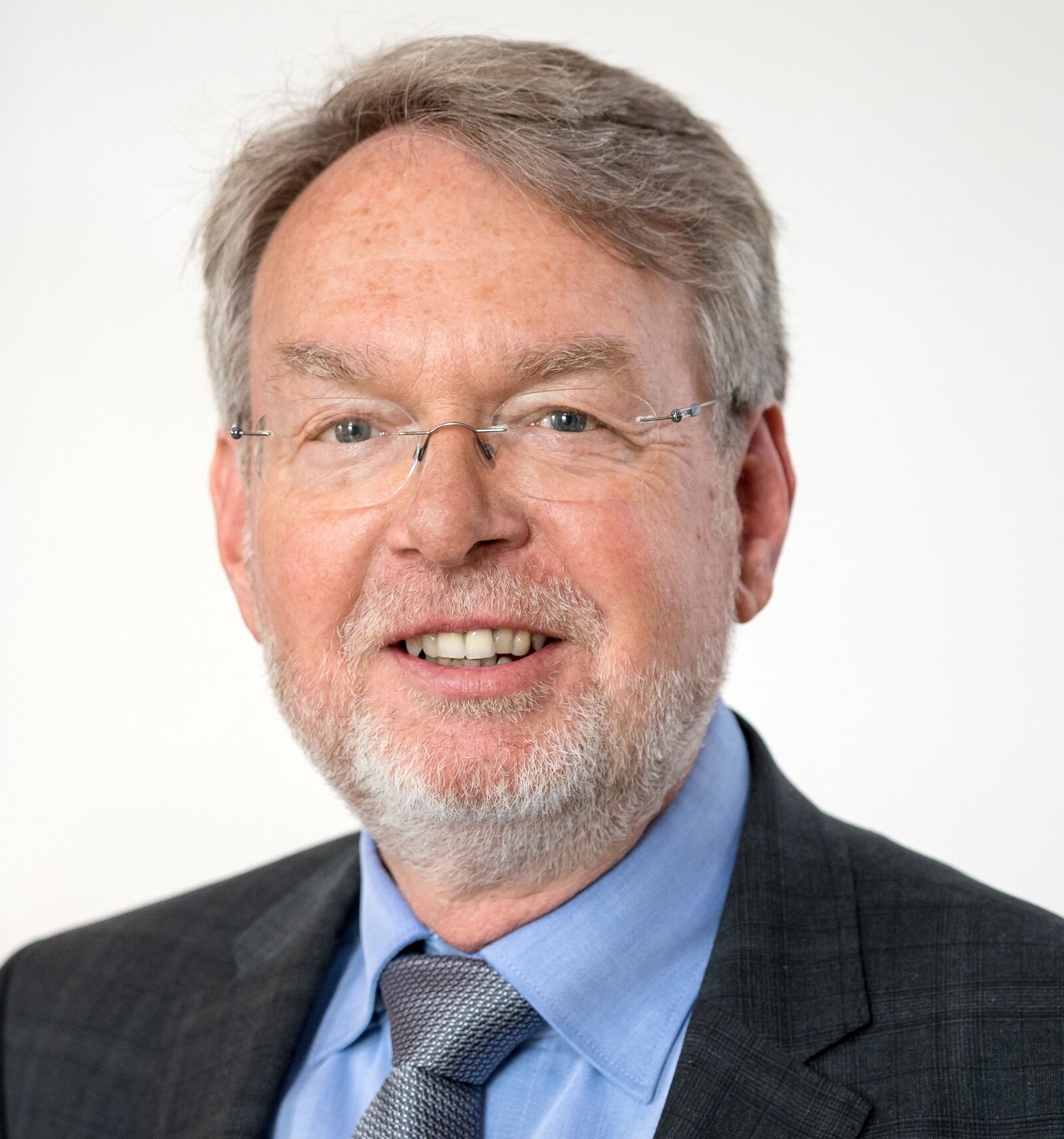 Ein Mann mit Brille und Bart lächelt in die Kamera.Er trägt eine Anzugjacke und Krawatte.