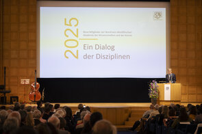 Ein Mann steht an einem Rednerpult. Im Hintergrund ist der Titel der Veranstaltung eingeblendet: Ein Dialog der Disziplinen.