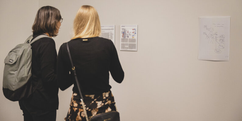 Zwei Frauen schauen sich einen gedruckten Text an, der an einer weißen Wand hängt.