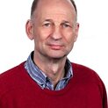 Ein Mann schaut freundlich in die Kamera. Er trägt ein kariertes Hemd unter einem roten Pullover.