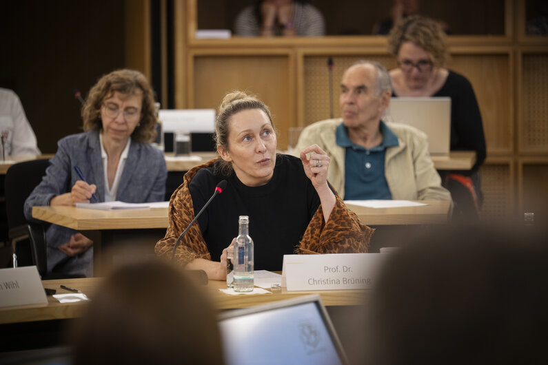 Eine Frau sitzt an einem Tisch. Auf einem Schild steht ihr Name "Prof. Dr. Christina Brüning".