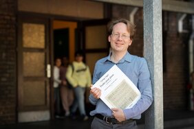 Porträtfoto Professor Dr. Tobias Bleek. Professor Bleek lehnt an einer Säule und hält die Noten eines Klavierstückes von György Ligeti in den Händen.