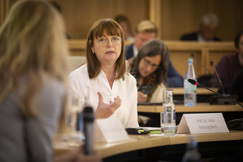 Eine Frau sitzt an einem Tisch. Auf einem Schild steht ihr Name "Prof. Dr. Viola Hartung-Beck".