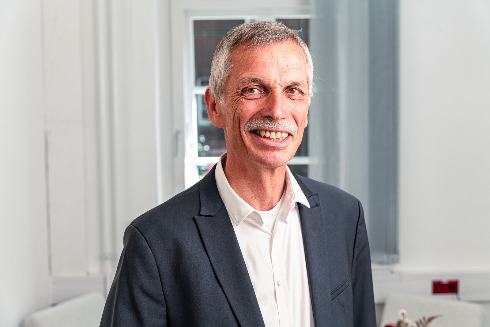 Ein Mann mit Oberlippenbart trägt eine Anzugjacke. Er steht vor einem Fenster und lächelt in die Kamera.