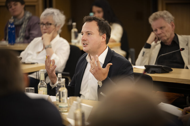 Ein Mann sitzt an einem Tisch. Er gestikuliert mit den Händen.