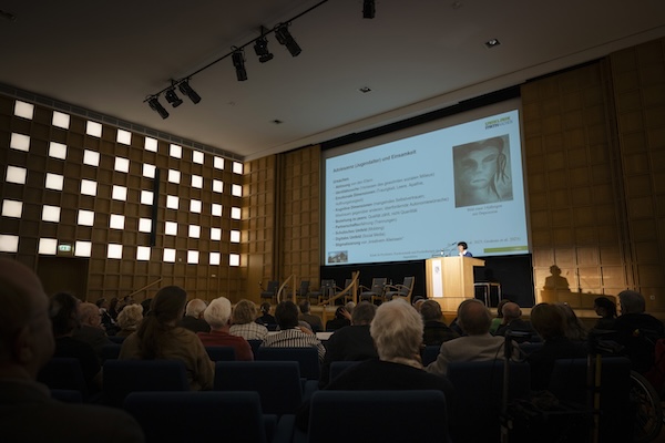 Ein gut gefüllter Veranstaltungssaal. Eine Frau hält einen Vortrag.