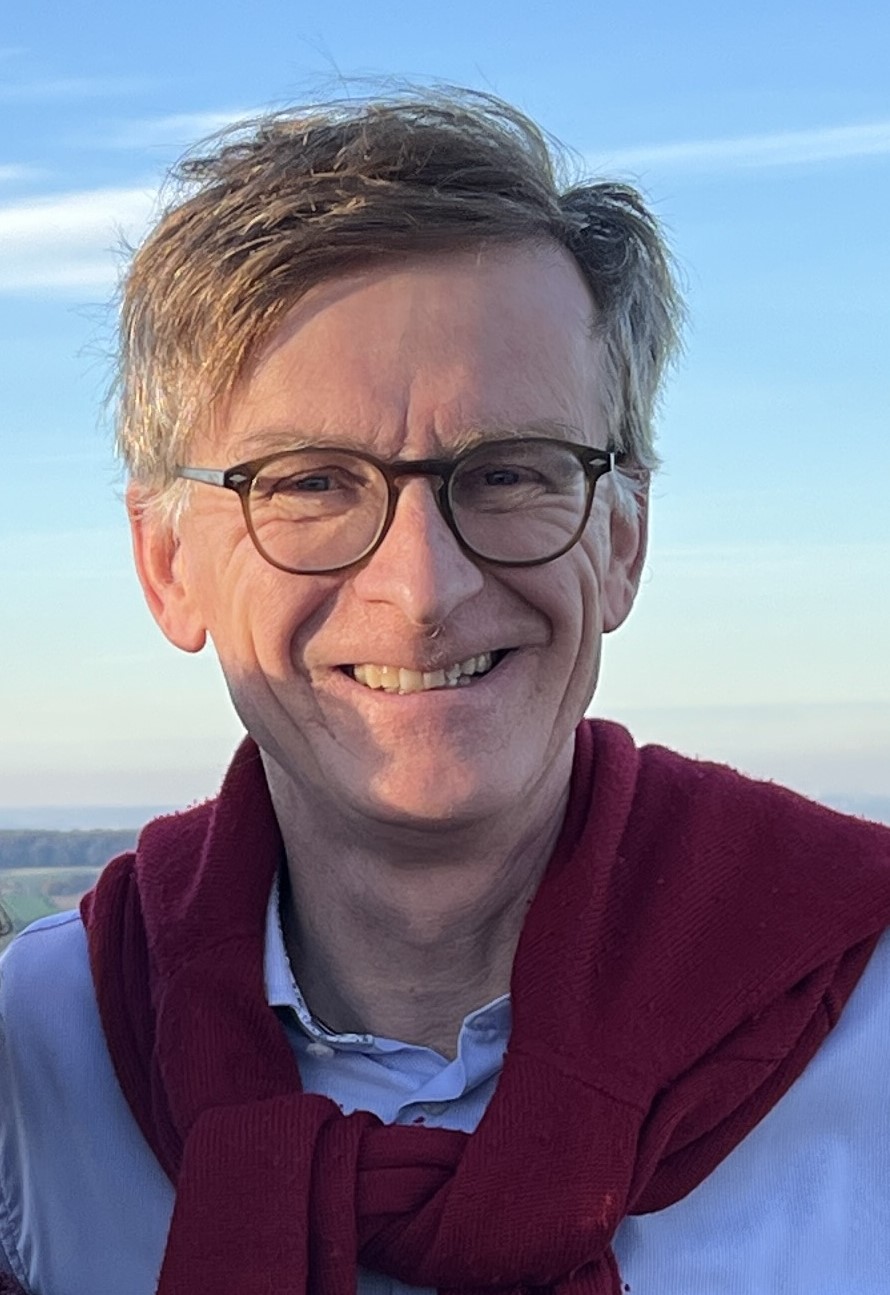 Ein Mann mit Brille und einem Pullover um die Schultern steht draueßen unter blauem Himmel. Er lächelt strahlend in die Kamera.