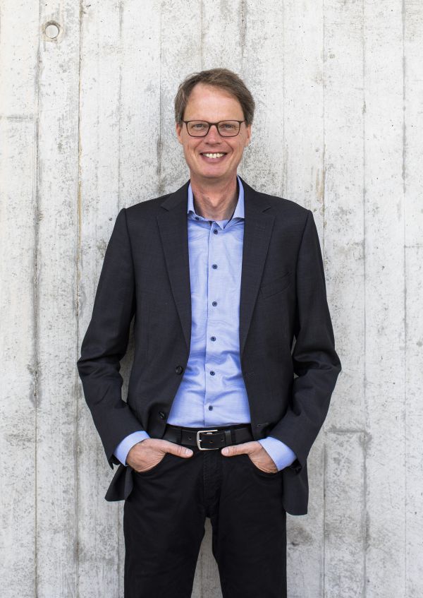 Ein Mann steht vor einer weißen Wand. Er trägt eine Anzugjacke und hat die Hände in den Hosentaschen. 