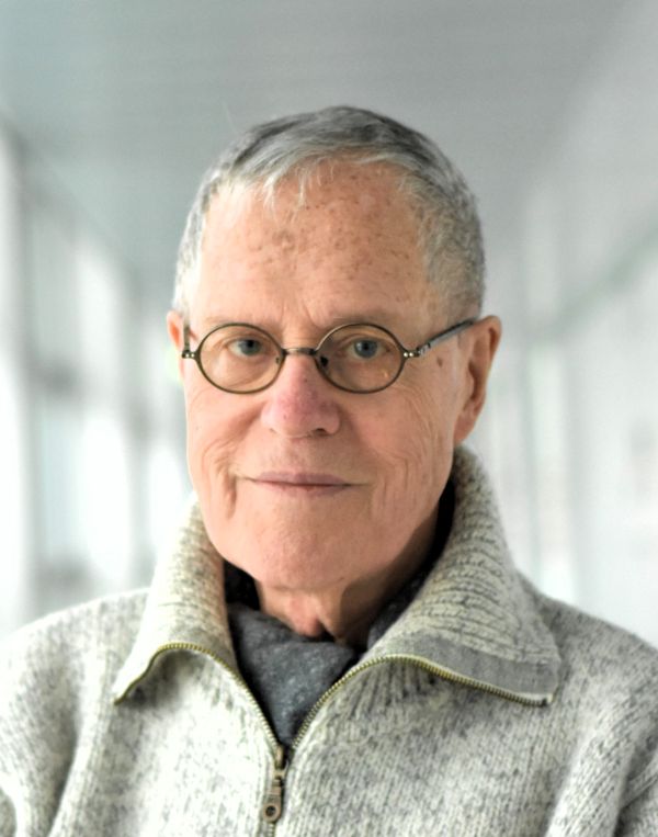 Ein Mann mit Brille trägt eine Strickjacke und einen Schal. Er lächelt in die Kamera.