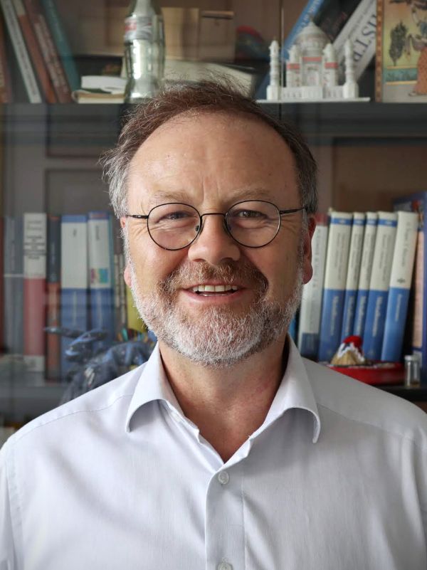 Ein Mann mit Brille und Bart steht vor einem Bücherregal und lächelt.