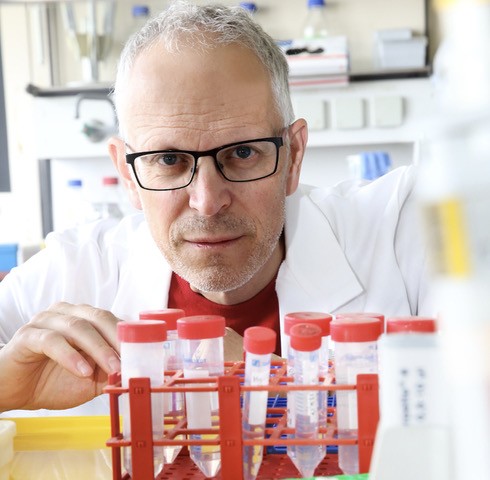 Ein Mann mit Brille befindet sich in einem Labor. Hinter einem Kasten mit Ampullen schaut er in die Kamera.