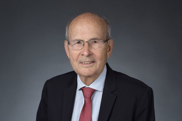 Ein Mann mit Brille trägt eine Krawatte und eine ANzugkacke. Er lächelt in die Kamera.