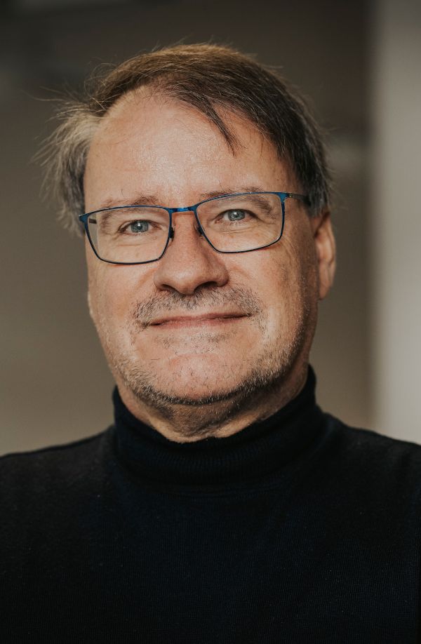 Ein Mann mit Brille lächelt in die Kamera. Er trägt einen schwarzen Rollkragenpullover.