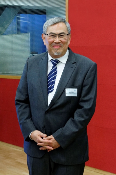 Ein Mann mit Brille steht lächelnd vor einer roten Wand. Er hat seine Hände vor dem Körper verschränkt.