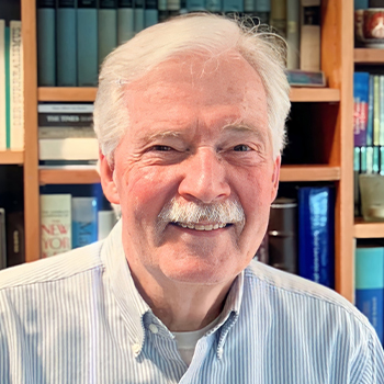 Ein Mann mit Oberlippenbart steht vor einem Bücherregal lund ächelt in die Kamera.