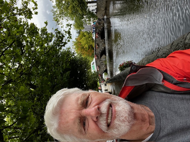 Ein Mann mit Bart steht unter einem Baum an einem Kanal. Im Hintergrund ist eine Brücke.