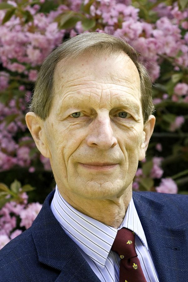 Ein Mann mit Anzug und Schlips steht vor einem blühenden Strauch und lächelt in die Kamera.